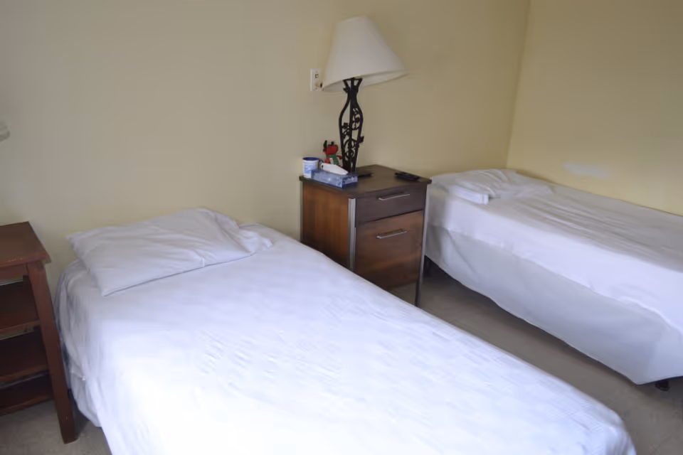 Two single beds with white bedding separated by a wooden nightstand and lamp in a simple room.