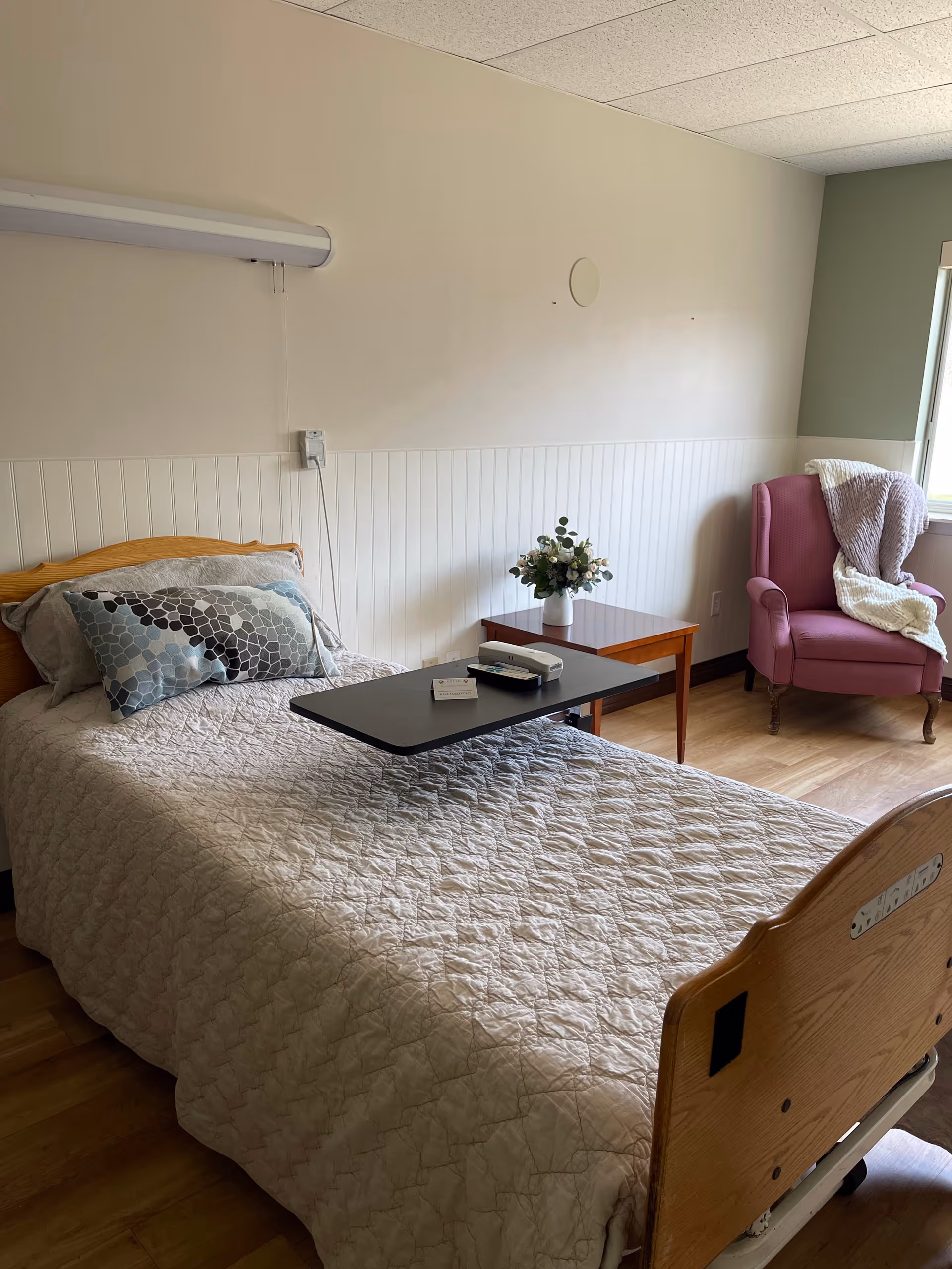 A neatly made single hospital-style bed with a quilted beige bedspread and a decorative pillow with a hexagonal pattern. There is a black overbed table with a remote control and a small card on it. Next to the bed is a wooden side table with a vase of flowers. In the corner near a window, there is a pink upholstered armchair with a white knitted blanket draped over it. The room has light-colored walls with white wainscoting and a wooden floor.