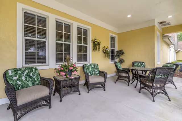Covered outdoor patio with wicker chairs and a table along a yellow wall under windows decorated with plants.