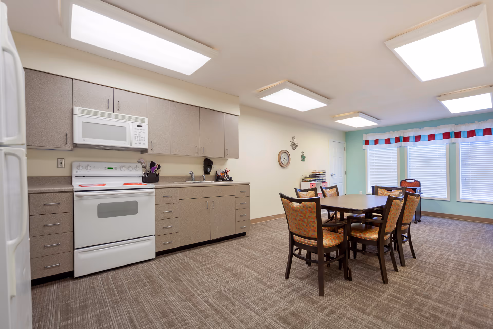 A bright kitchen and dining area in an assisted living facility featuring beige cabinets, a white stove with a microwave above it, a sink, and a refrigerator. There is a wooden dining table with six patterned chairs on a carpeted floor. The room has large windows with blinds and a colorful valance, and the walls are painted in light colors with a clock and wall decorations.