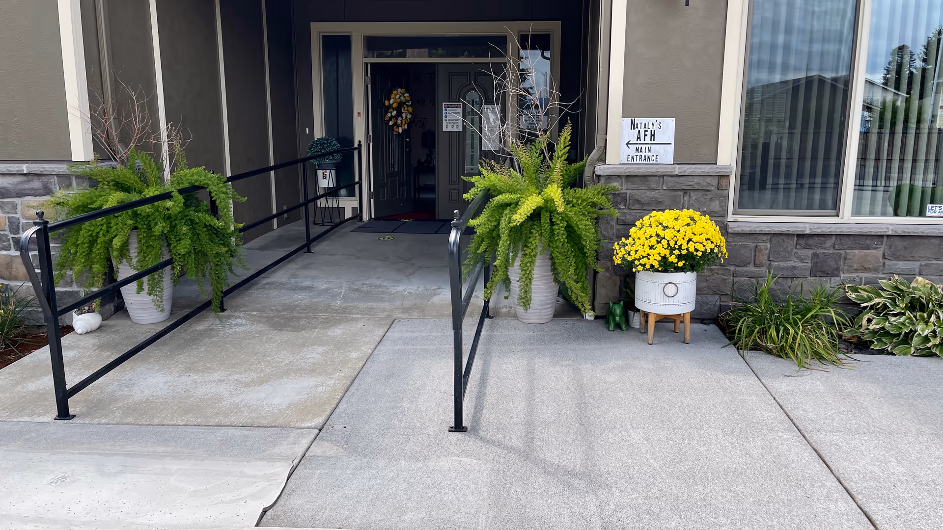Entrance to Nataly's Adult Family Home with a concrete ramp and black metal railings on both sides. Two large potted green plants flank the entrance, and a pot of yellow flowers is placed near the wall. A sign on the wall reads 'Nataly's AFH Main Entrance'.