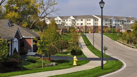 A paved road curves through a landscaped area with green grass, trees, and shrubs. On the left side, there are residential-style buildings with brick and siding exteriors. In the background, a larger multi-story building with balconies is visible. A streetlamp and a yellow fire hydrant are also present along the sidewalk.