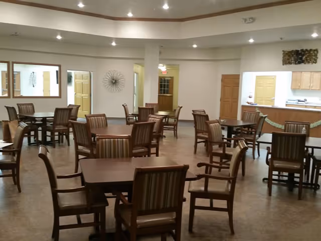 A spacious dining area with multiple wooden tables and chairs arranged neatly. The room has a high ceiling with recessed lighting and light-colored walls. There is a kitchen area visible in the background with wooden cabinets and a countertop.