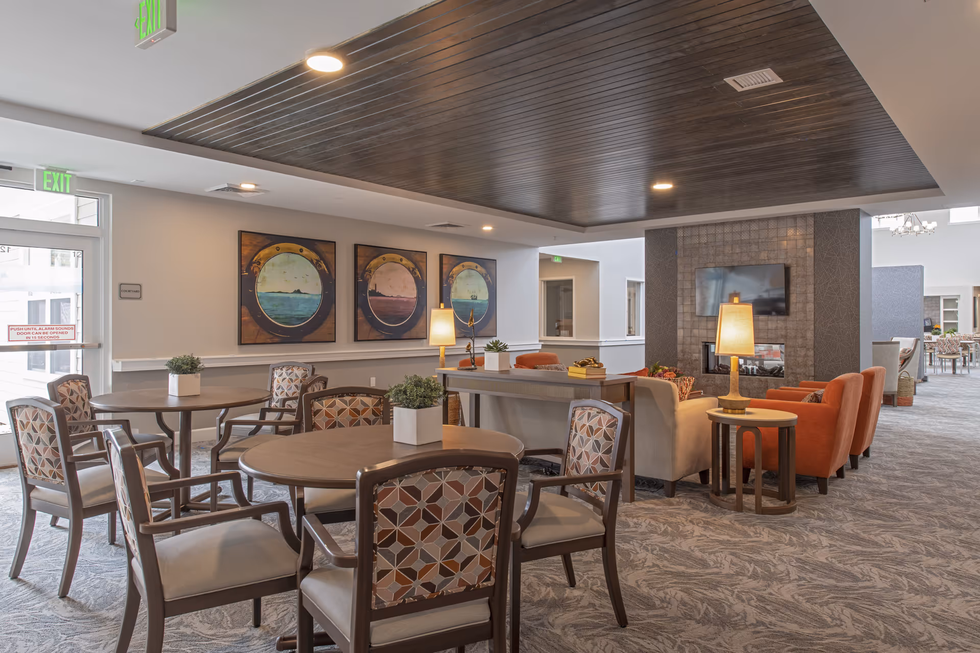 Well-lit common area with round dining tables and patterned chairs adjacent to a cozy seating area with couches, armchairs, lamps, and a wall-mounted TV.