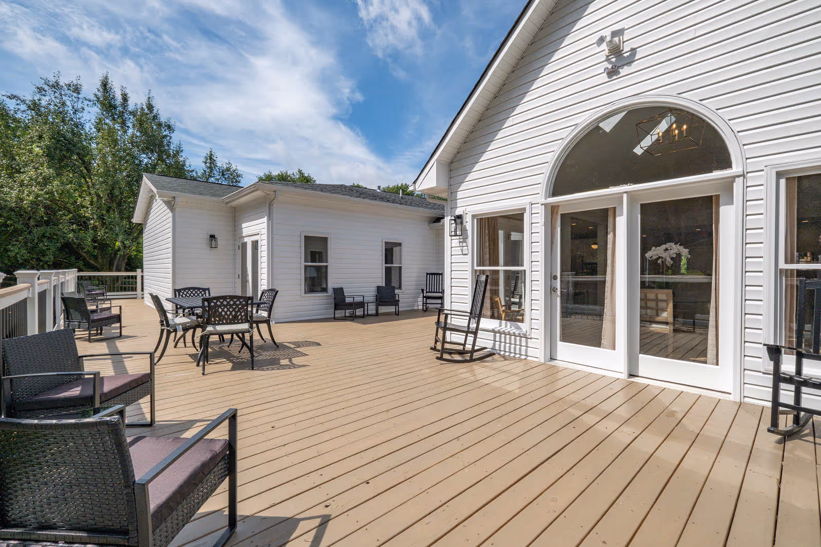 Spacious outdoor wooden deck area with multiple seating arrangements including chairs and tables. The deck is attached to a white building with large glass doors and windows, under a partly cloudy blue sky.