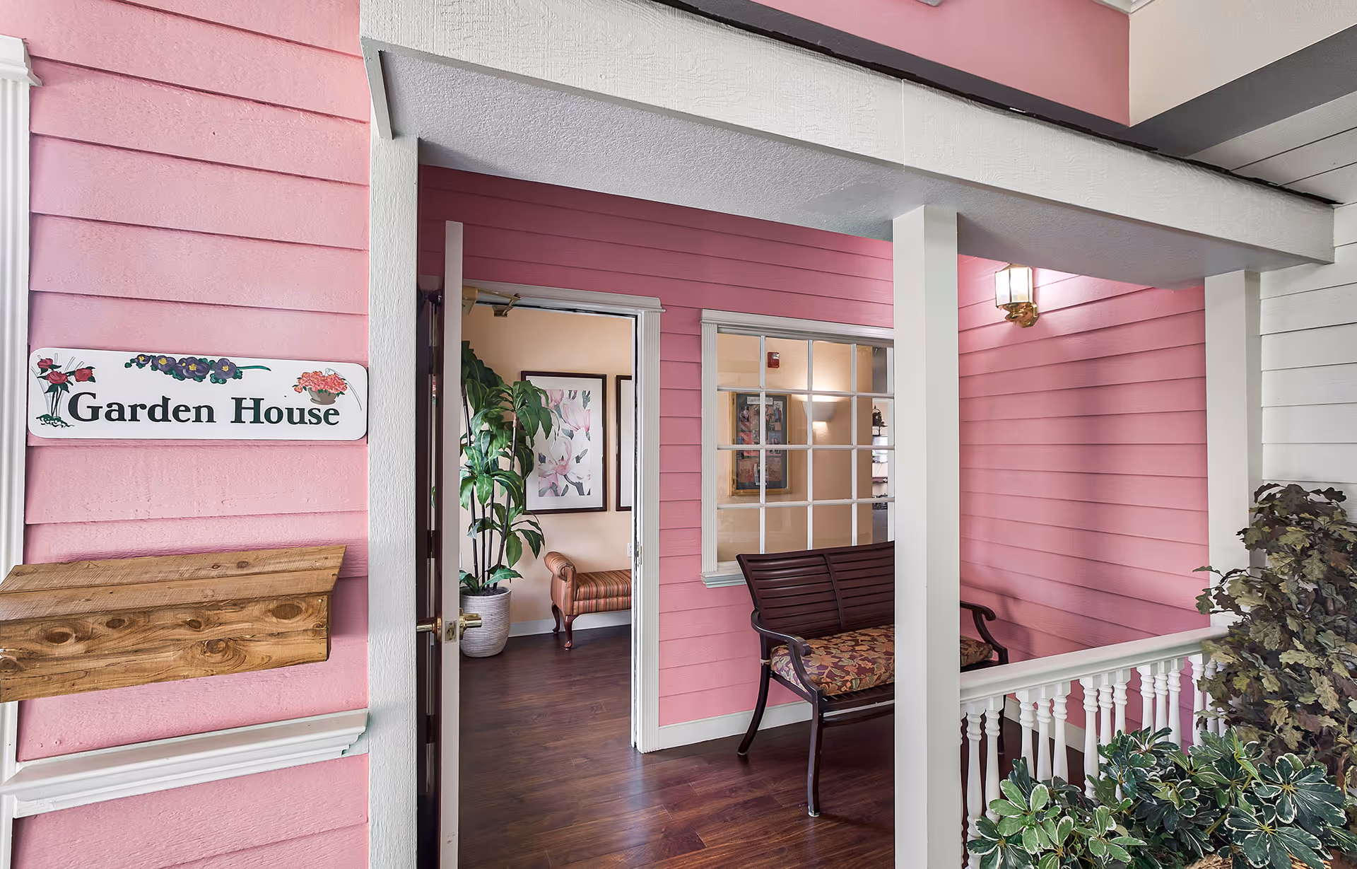 Entrance to the Garden House at Juniper Village at Aurora with pink exterior walls, a wooden bench with floral cushion, potted plants, and an open door leading to an interior room with framed floral artwork and a striped armchair.