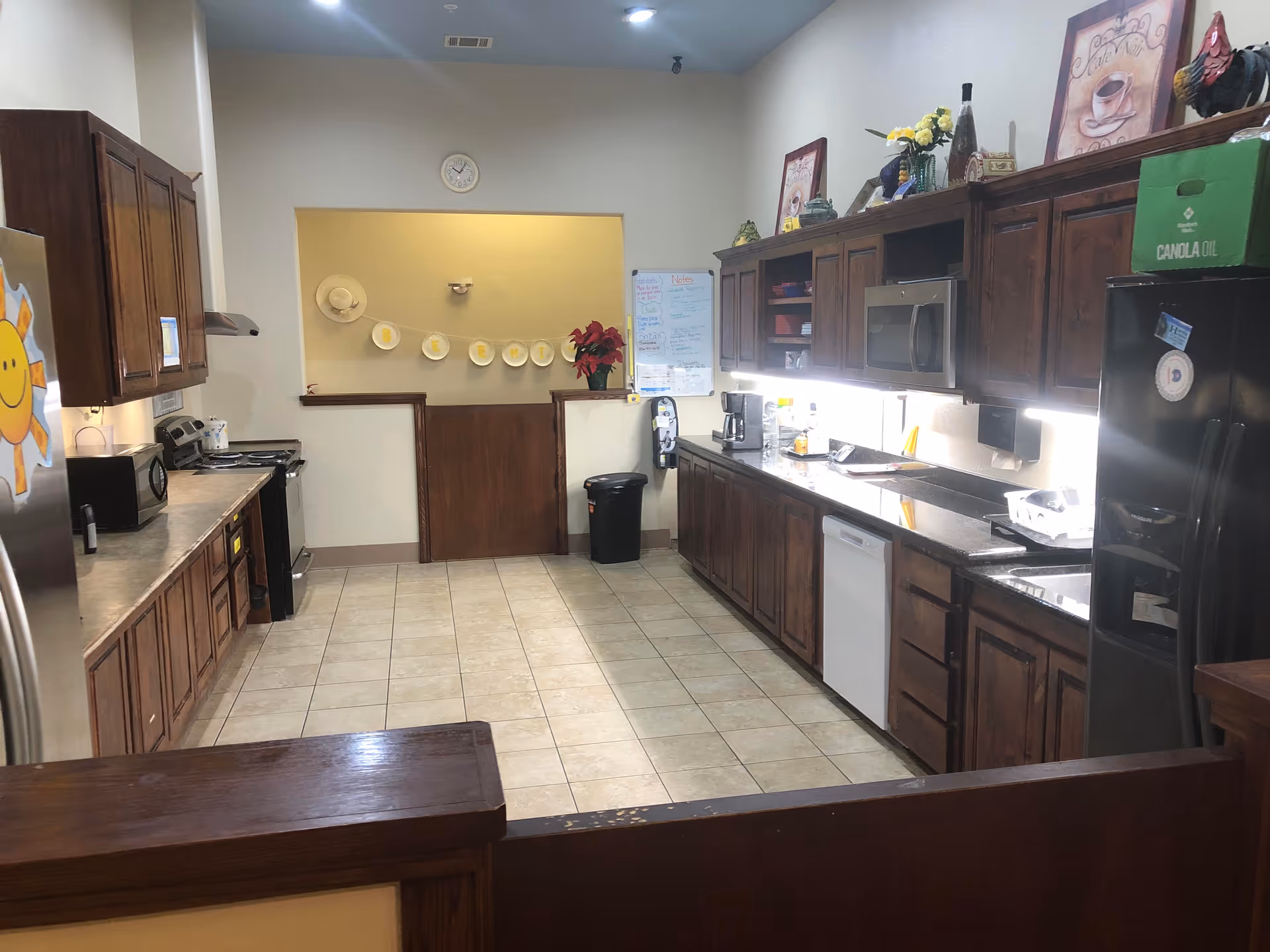 Communal kitchen with long countertops, wooden cabinets, appliances, and a tiled floor in a senior living facility.
