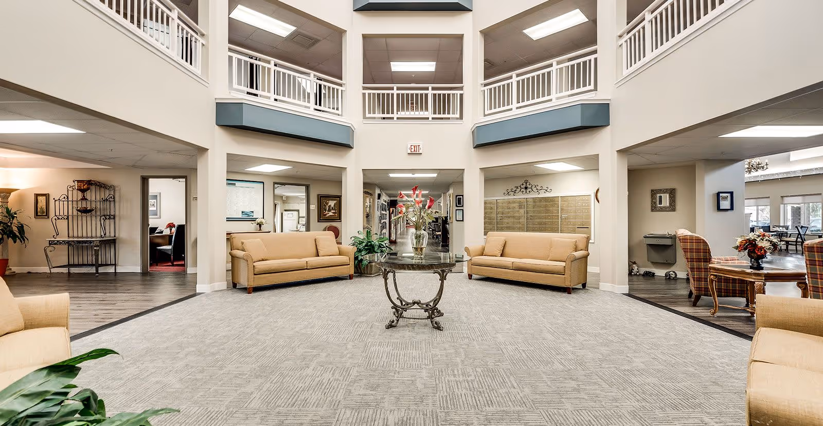 Spacious and well-lit common area in a senior living facility with beige sofas arranged around a central glass table holding a vase with flowers. The area features high ceilings with a second-floor balcony overlooking the space. There are plants and decorative elements, with additional seating and tables visible in adjoining rooms.