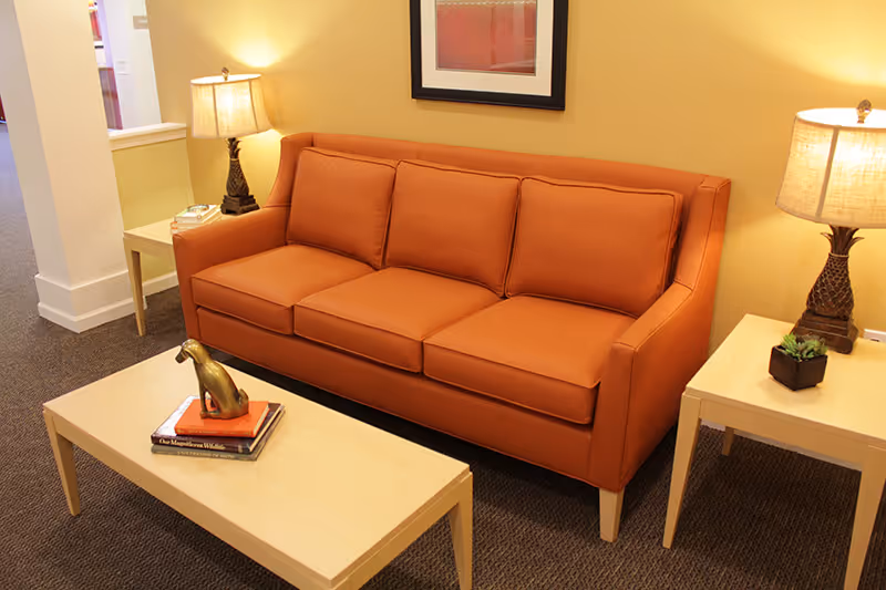 A cozy living room area featuring an orange three-seat sofa flanked by two wooden side tables, each with a lamp. A wooden coffee table in front of the sofa holds a small decorative sculpture and a couple of books. The walls are painted a warm yellow, and a framed picture hangs above the sofa.
