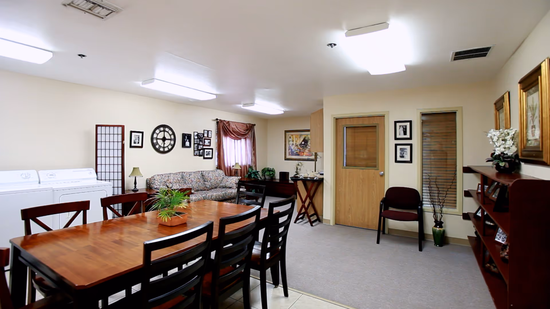 A communal room with a wooden dining table and six chairs, a floral patterned couch, a washing machine and dryer, a wooden bookshelf with framed pictures and decorations, a door with a window, and framed artwork on the walls. The room is well-lit with ceiling lights and has a carpeted floor area and tiled floor near the dining table.