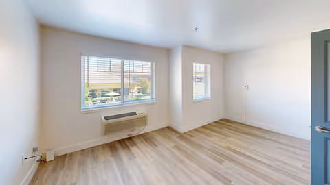 Empty bright room with wood-look flooring, two windows and a wall-mounted air conditioning/heater unit beneath one window.