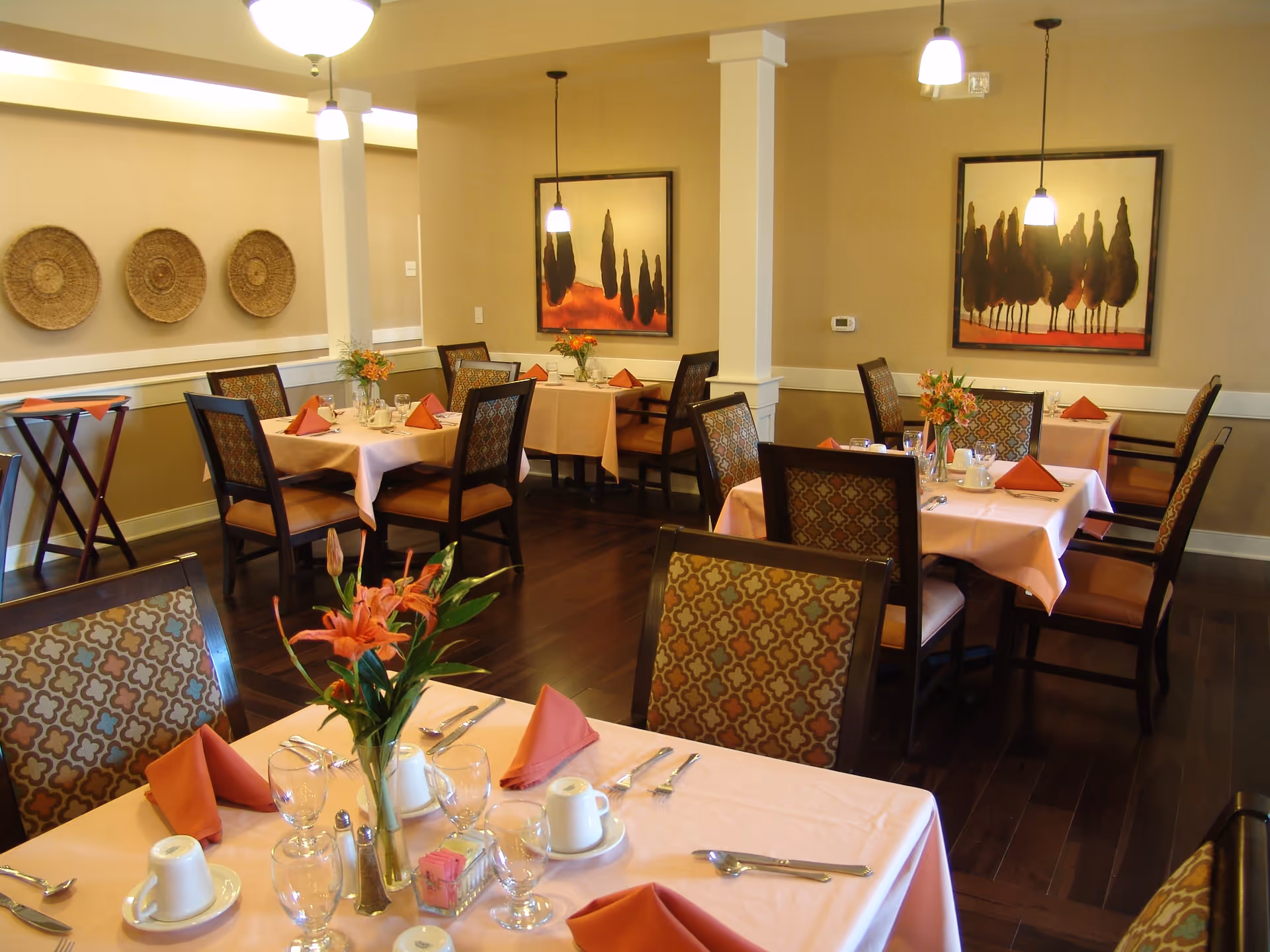 A dining room with several tables covered in light pink tablecloths, each set with orange folded napkins, glassware, and white cups. The chairs have patterned upholstery. The room has wooden flooring, beige walls, decorative wall art, and hanging pendant lights. There are also flower arrangements on the tables.