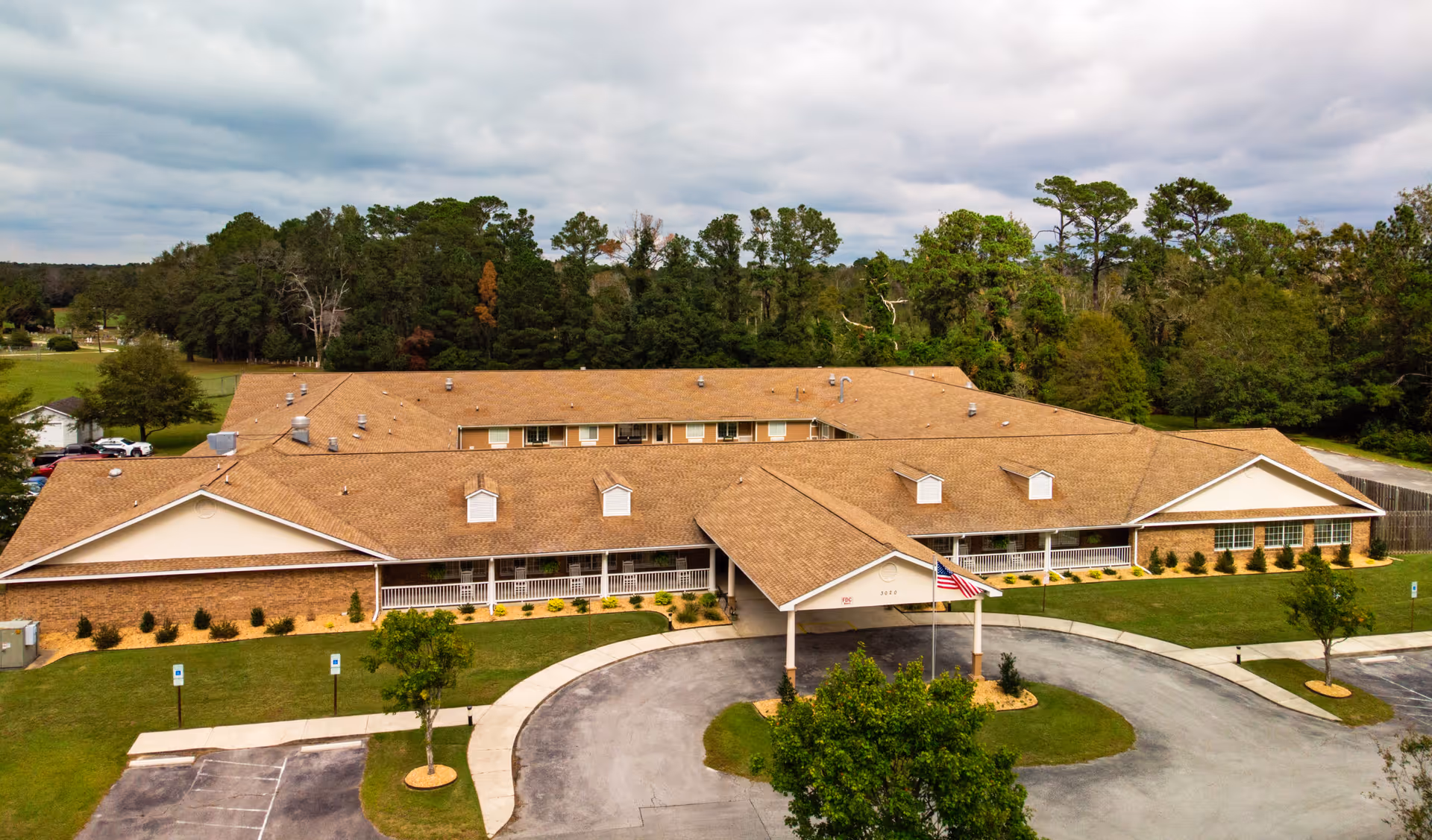 Aerial view of a single-story senior living facility building with a tan roof and brick exterior, surrounded by green lawns and trees under a cloudy sky. There is a circular driveway with an American flag near the entrance.