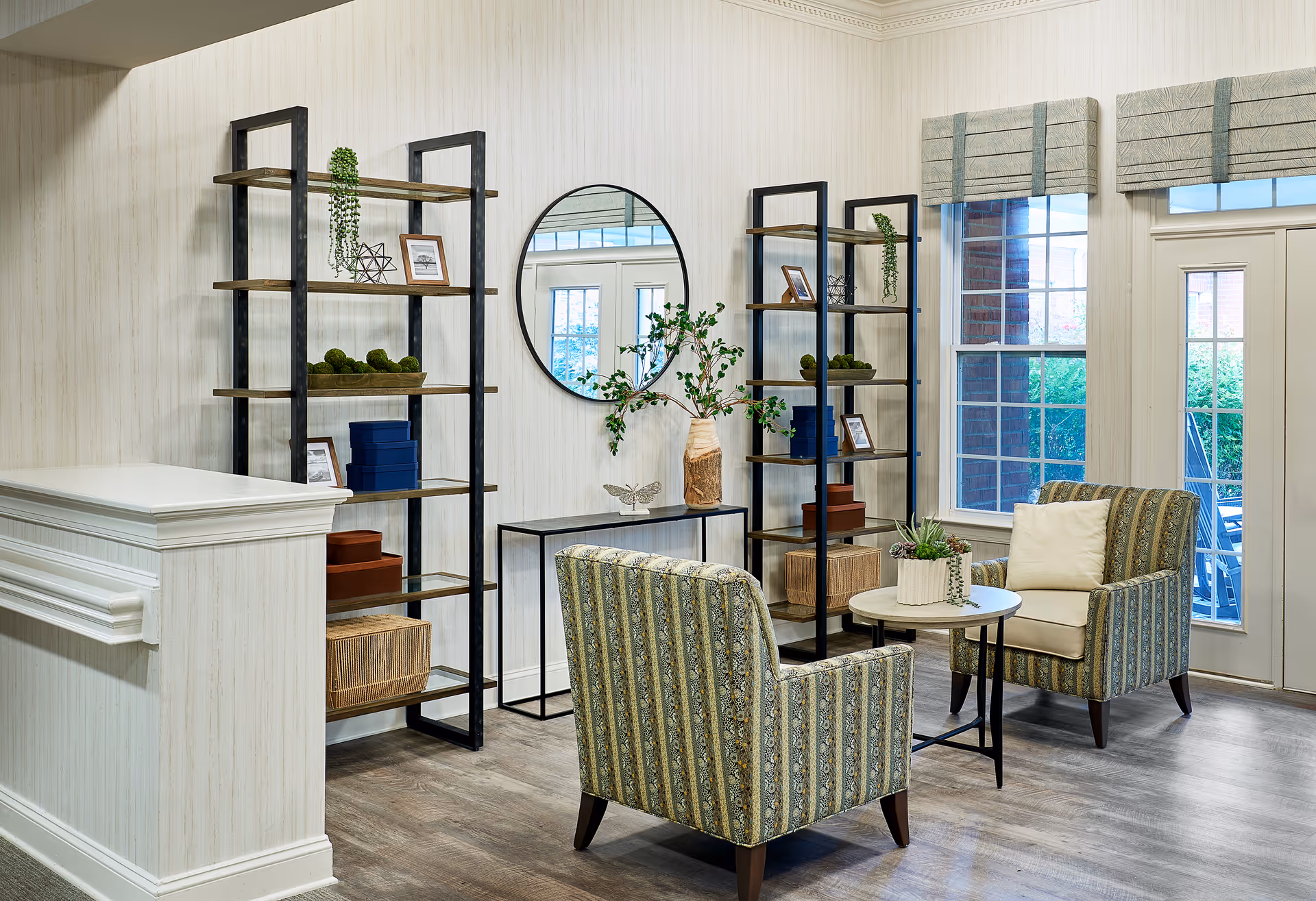 A cozy sitting area in a senior living facility featuring two patterned armchairs with cushions, a small round table with a potted plant, two tall black metal and wood shelving units with decorative items, a round wall mirror, and large windows with beige Roman shades letting in natural light.