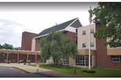 Front view of a three-story brick and beige senior healthcare building with a covered drop-off entrance, driveway, and landscaping.