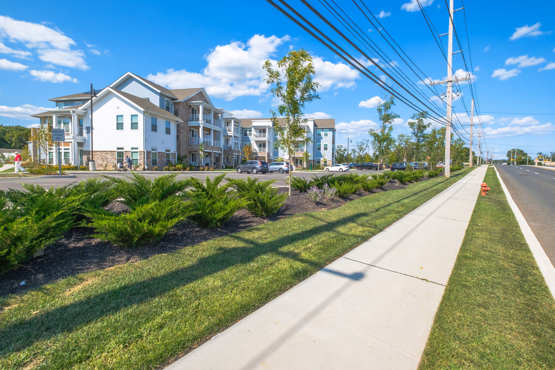 Exterior view of a senior living facility building with multiple floors, balconies, and a parking lot. The scene includes a sidewalk, green grass, shrubs, small trees, power lines, and a clear blue sky with scattered clouds.