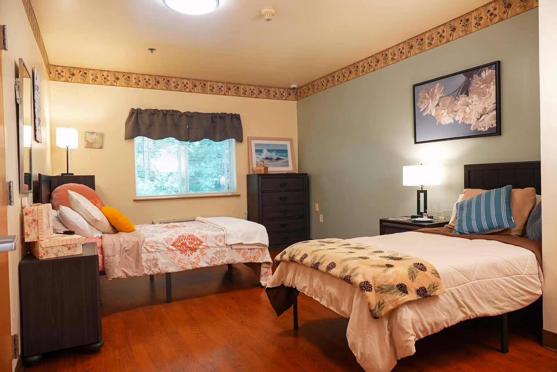 A cozy bedroom with two beds, one twin and one full, each with colorful bedding and pillows. The room has wooden flooring, a window with blinds and a valance, a dresser with a framed picture on top, and a nightstand with a lamp. The walls are painted in two tones with a floral border near the ceiling.