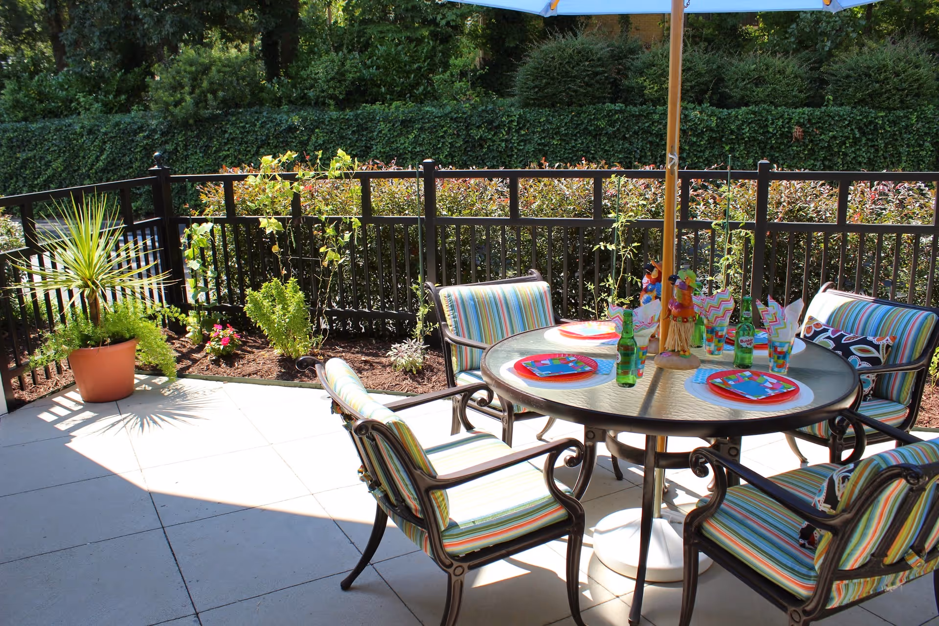 Outdoor patio area with a round glass table and four striped cushioned chairs. The table is set with colorful plates, napkins, and green glass bottles. A large blue umbrella provides shade, and there are potted plants and a black metal fence surrounding the patio. Greenery and bushes are visible in the background.