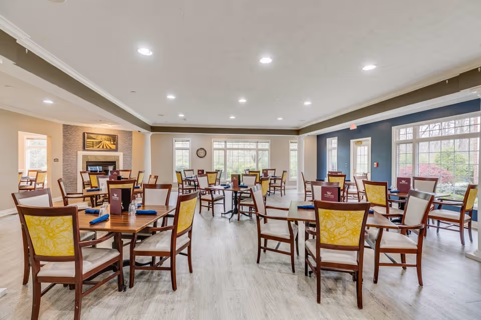 A bright and spacious dining room with multiple wooden tables and chairs arranged neatly. Each table has menus and blue napkins placed on it. Large windows allow natural light to fill the room, and there is a fireplace with a framed picture above it on one wall. The flooring is light-colored wood, and the ceiling has recessed lighting.