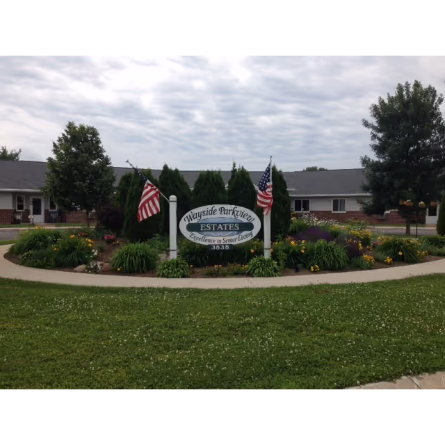 Outdoor view of Wayside Parkview Estates senior living facility sign surrounded by landscaped greenery and flowers, with two American flags on either side. The building and trees are visible in the background under a cloudy sky.