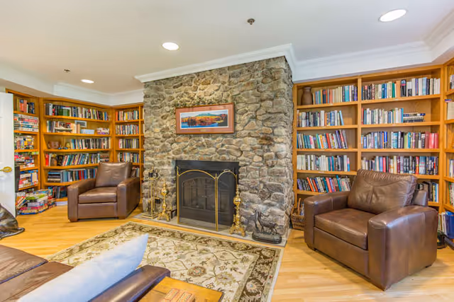 Cozy living room with a stone fireplace in the center, flanked by built-in wooden bookshelves filled with books. Two brown leather armchairs are positioned on either side of the fireplace, and a patterned area rug covers the wooden floor in front of it. A framed landscape painting hangs above the fireplace.