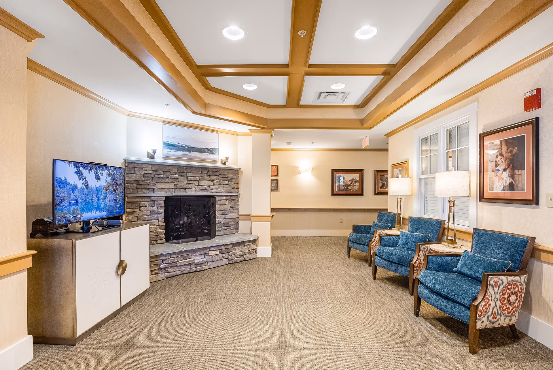 A cozy communal lounge with a stone fireplace, television and a row of blue upholstered chairs under a coffered ceiling.
