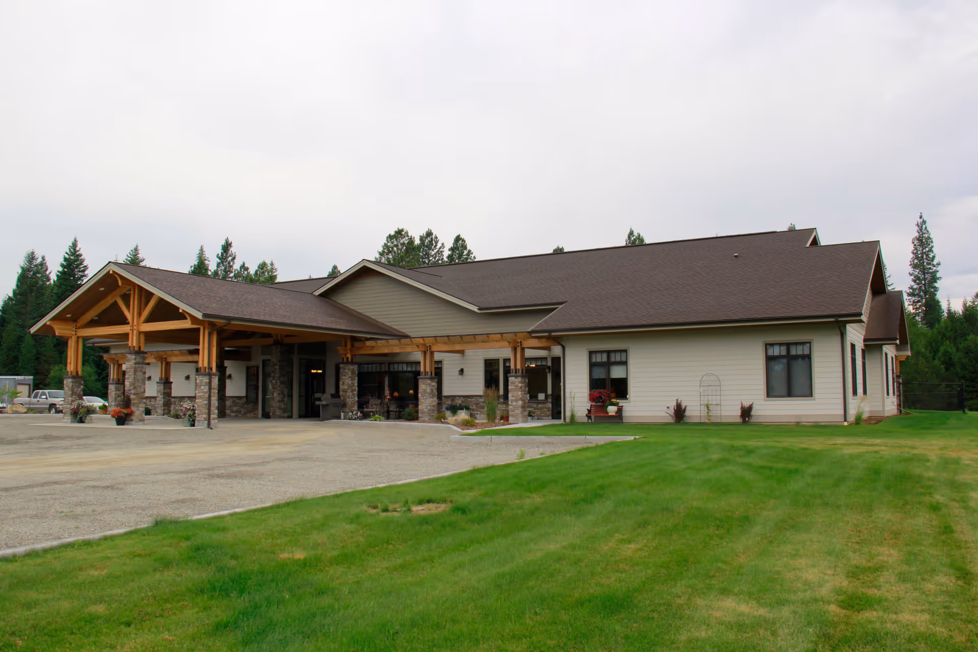 Exterior view of a single-story assisted living facility building with a large covered entrance supported by stone and wooden pillars. The building is surrounded by a well-maintained green lawn and trees in the background under a cloudy sky.