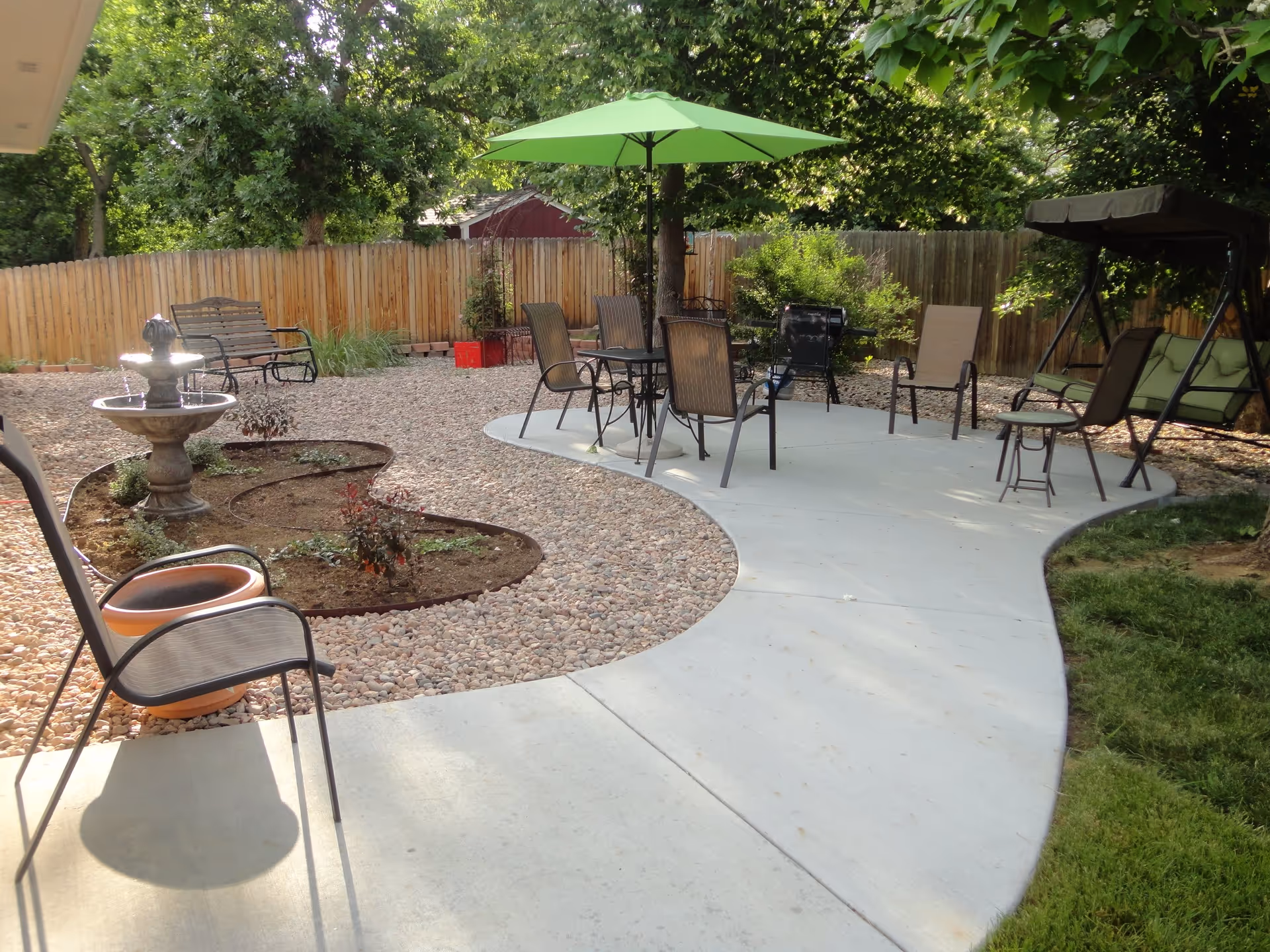 Outdoor patio area with a curved concrete walkway surrounded by gravel and small garden beds. There is a round table with four chairs under a green umbrella, a bench, a swing with cushions, a small side table, and a water fountain. The area is enclosed by a wooden fence and shaded by trees.