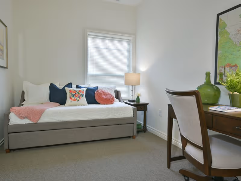 A cozy bedroom with a gray daybed adorned with white, navy blue, floral, and pink pillows, a pink throw blanket, a wooden side table with a lamp, and a wooden desk with a chair. A large window with blinds allows natural light into the room, and a framed map hangs on the wall above the desk.