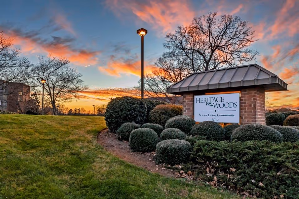 A landscaped outdoor area at sunset with a brick sign that reads 'Heritage Woods Senior Living Community 3812' surrounded by bushes and grass, with a streetlamp and trees in the background under a colorful sky.