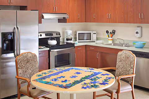 A kitchen area with wooden cabinets, a stainless steel refrigerator, oven, microwave, and dishwasher. A round table in the center has a partially completed jigsaw puzzle on it, surrounded by two upholstered chairs with wooden armrests. Various kitchen utensils and a coffee maker are on the countertop.