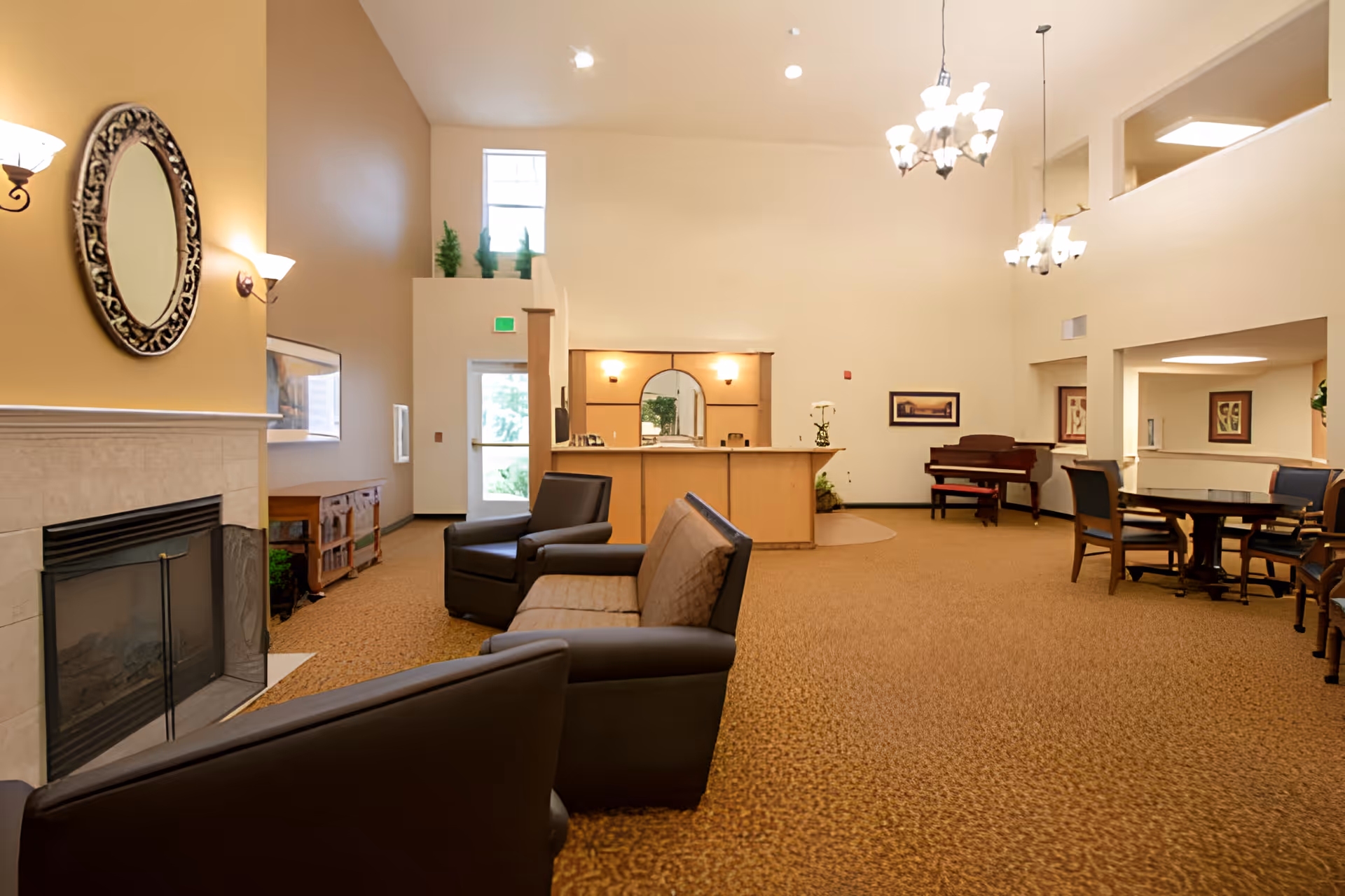 A spacious senior living facility common area with beige walls and carpet. The room features a fireplace with a decorative round mirror above it, several armchairs and a sofa arranged for seating, a reception desk with a mirror behind it, a piano in the corner, and a dining table with chairs. The area is well-lit with wall sconces and chandeliers, and there are framed pictures on the walls.