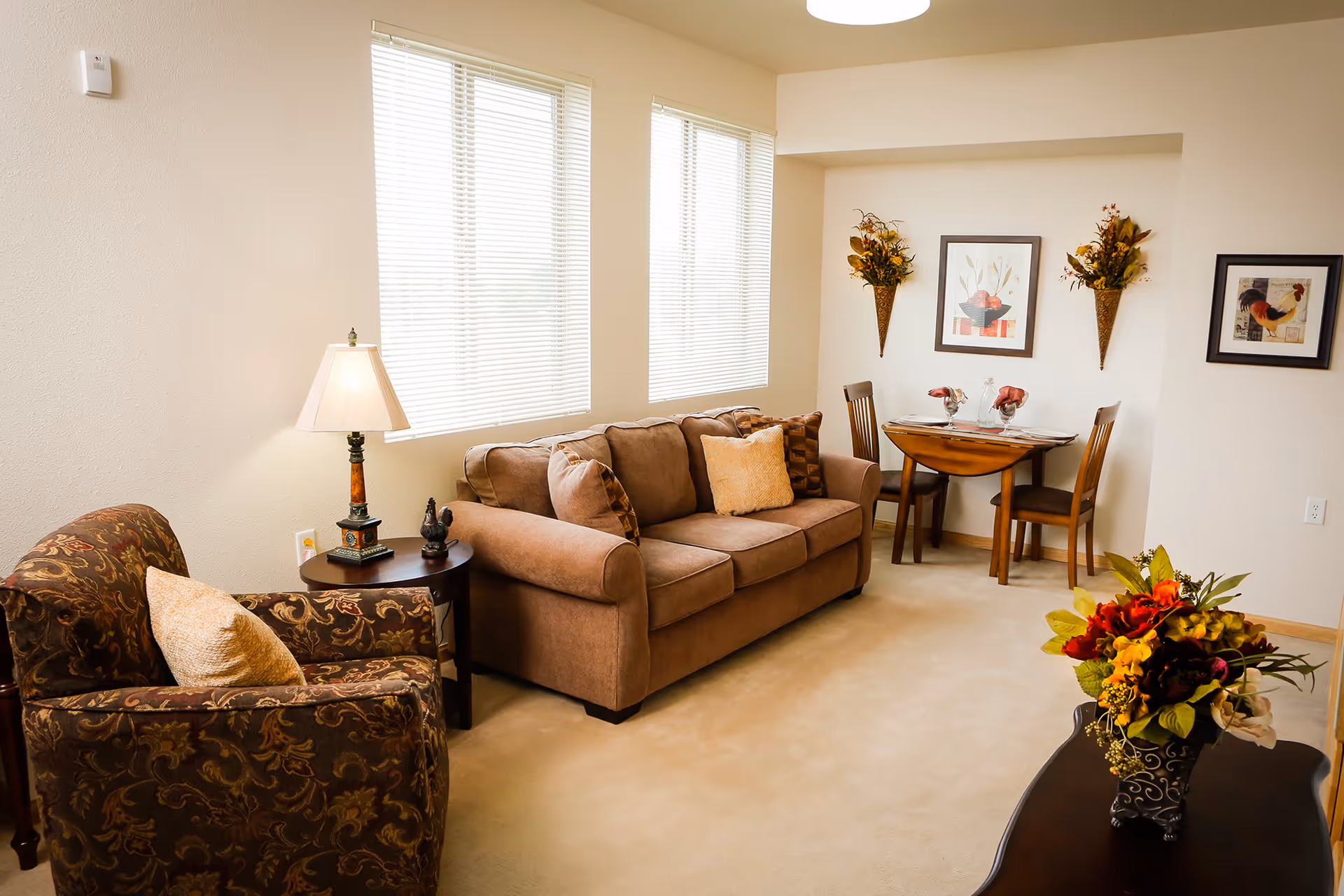 A cozy living room area with a brown sofa adorned with multiple cushions, a patterned armchair with a beige pillow, and a small round wooden side table with a lamp and decorative items. In the background, there is a small wooden dining table set for two with framed artwork and wall-mounted floral arrangements on the walls. A coffee table with a floral arrangement is in the foreground.