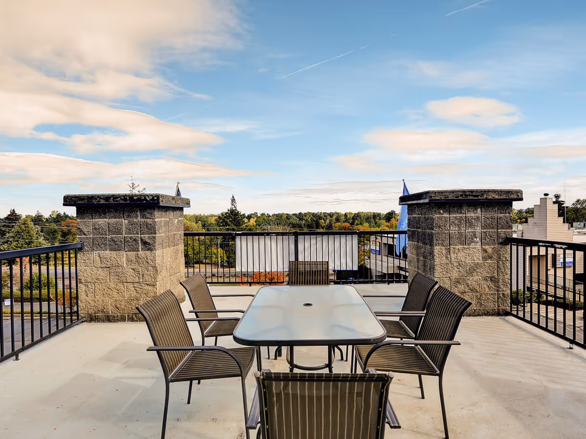 Outdoor patio area with a glass-top table and six metal chairs arranged around it, surrounded by black metal railings and two large stone pillars, overlooking a view of trees and buildings under a partly cloudy sky.
