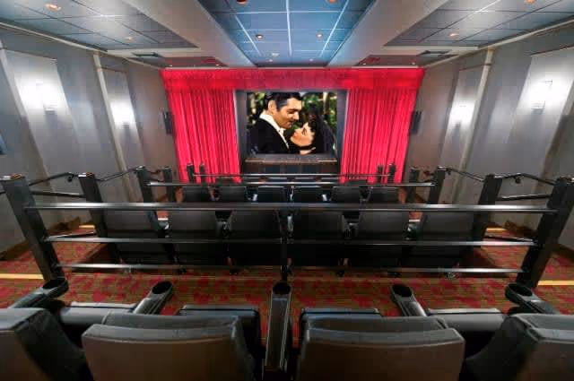 Interior view of a small movie theater with rows of cushioned seats facing a screen displaying a romantic scene of a man and woman. The theater has red curtains around the screen and soft lighting on the walls.