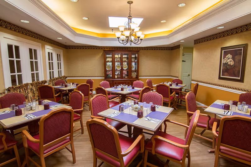 Dining room with multiple set tables and red upholstered chairs, a chandelier, china cabinet and framed artwork on the walls.