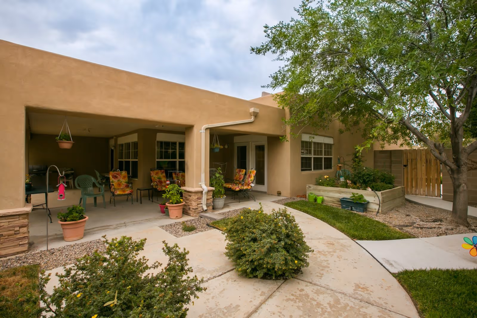 Outdoor patio area of a senior living facility with several chairs featuring floral cushions, potted plants, a small garden bed, and a tree. The building has a beige stucco exterior with large windows and a covered seating area.