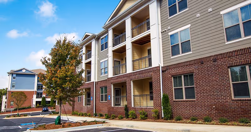Exterior view of a multi-story residential building with brick and siding facade, balconies, and a parking lot with a tree in the foreground under a blue sky with some clouds.