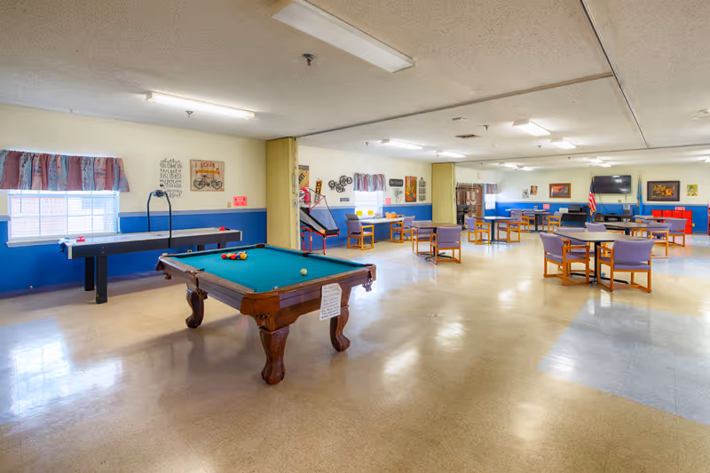 A spacious recreational room with a pool table and an air hockey table near the windows. Several tables with chairs are arranged throughout the room, and there are various decorations and framed pictures on the walls. The room has a tiled floor and fluorescent ceiling lights.