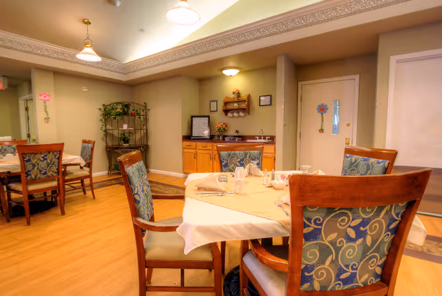 A dining area in a senior living facility with wooden chairs featuring patterned upholstery around tables covered with beige tablecloths. The room has warm lighting, a wooden floor, and a small cabinet with a sink and decorative items against the wall. There are flower decorations on the walls and a metal shelf with plants in the background.
