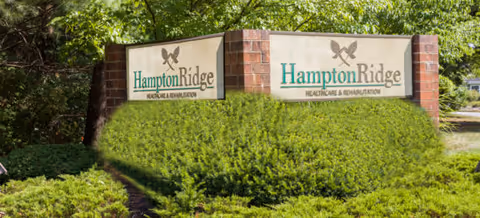Entrance sign reading 'Hampton Ridge Healthcare & Rehabilitation' mounted on brick pillars surrounded by shrubs and trees.