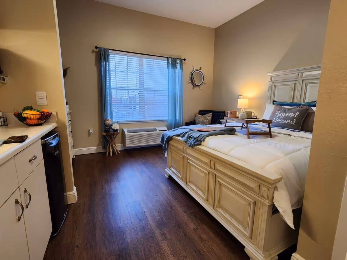 A cozy bedroom with a large bed featuring white bedding and pillows, one pillow has the text 'Beyond blessed'. There is a small wooden tray on the bed with a lamp and mugs on a side table next to a blue cushioned chair. A window with blue curtains and blinds lets in natural light. The room has wooden flooring and beige walls. On the left side, part of a kitchenette with white cabinets and a small black refrigerator is visible.