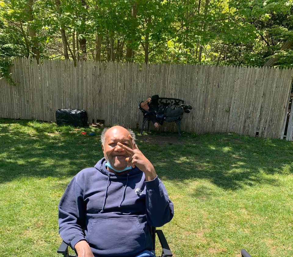Two people in a sunny backyard—one seated in a lawn chair near the camera making a hand gesture and another reclining on a bench by a wooden fence under trees.