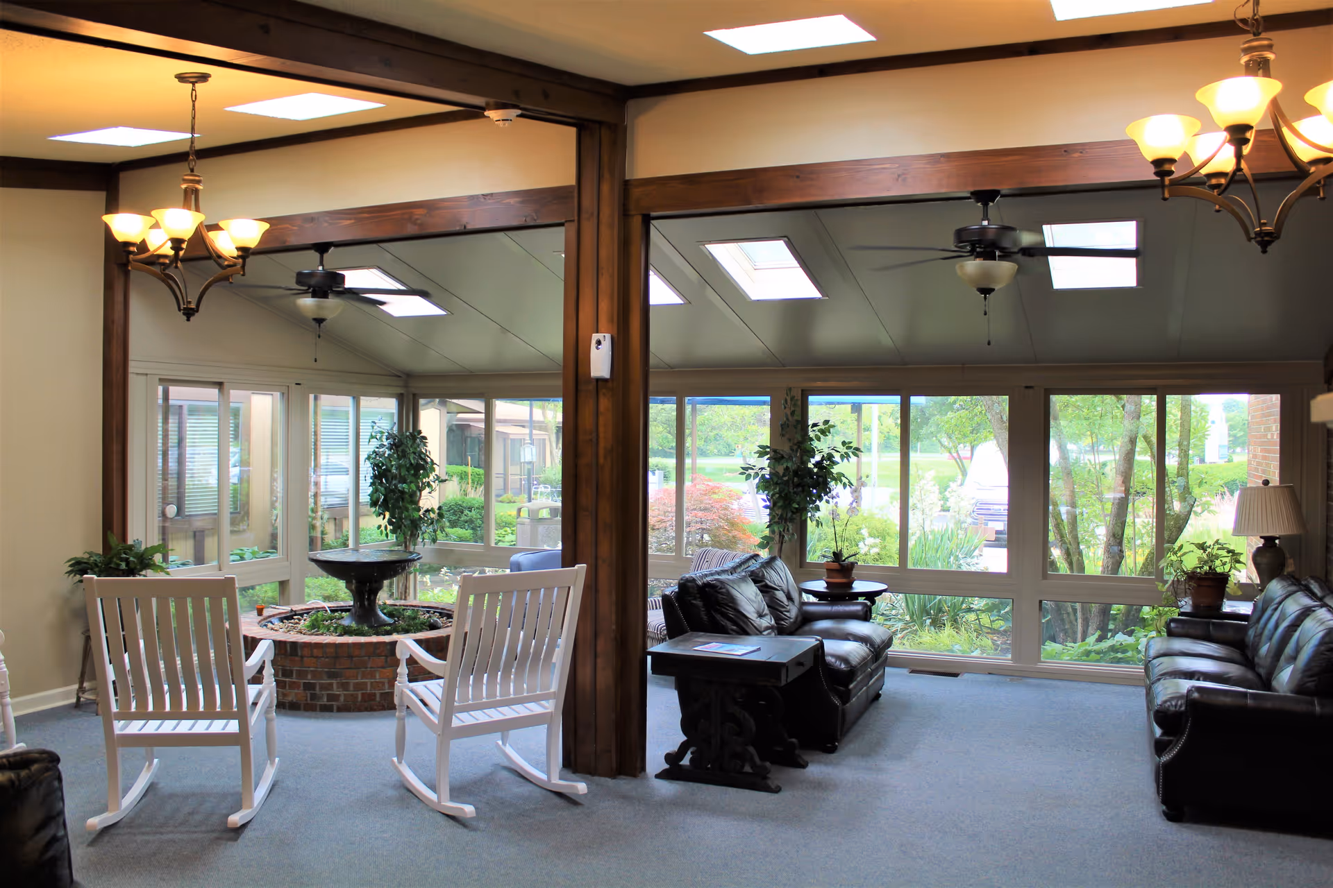 A bright and cozy indoor sitting area with large windows showing a garden outside. The room features two white rocking chairs facing a brick planter with a fountain, two black leather sofas, a small wooden side table, potted plants, ceiling fans, and warm overhead lighting.
