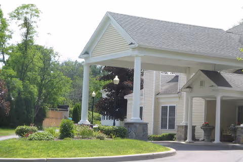 Entrance of a residential building with a covered driveway supported by white columns, surrounded by green grass, bushes, and trees under a clear sky.