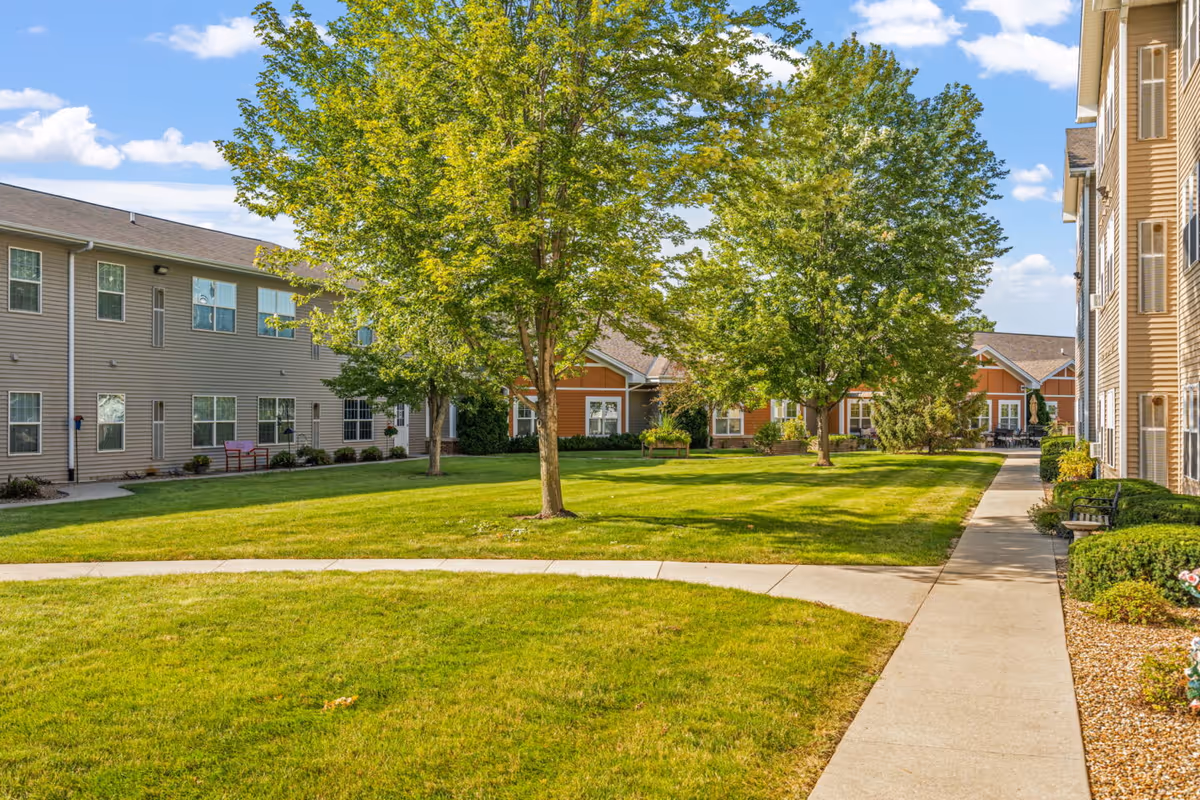 A well-maintained outdoor courtyard area with green grass, several trees, and a concrete walkway. Surrounding the courtyard are multi-story residential buildings with beige and orange siding under a partly cloudy blue sky.