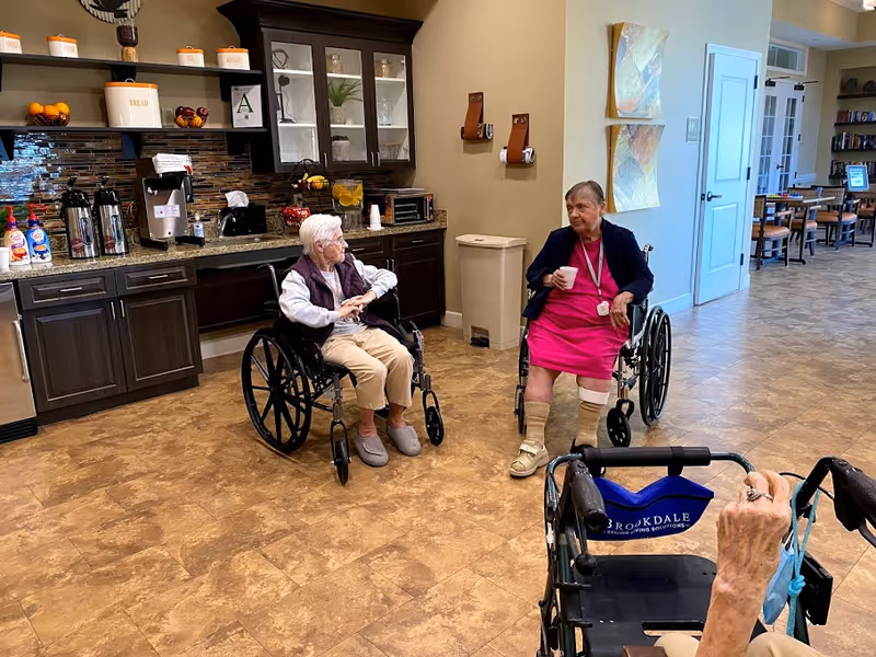 Two elderly women in wheelchairs are conversing in a common area with a kitchenette behind them. The kitchenette has dark wood cabinets, a coffee station with creamers, a toaster oven, and shelves with decorative items. The floor is a light brown tile, and there are two abstract paintings on the wall. A third person is partially visible in the foreground using a walker with a blue Brookdale Senior Living Solutions bag.