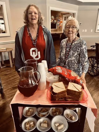 Two elderly women standing behind a table with a pitcher of iced tea, stacked disposable cups, a bag of Lay's potato chips, and a plate of stacked sandwiches. Below the table are several bowls containing sliced bananas and other fruit. The setting appears to be an indoor common area with wooden flooring and neutral-colored walls.