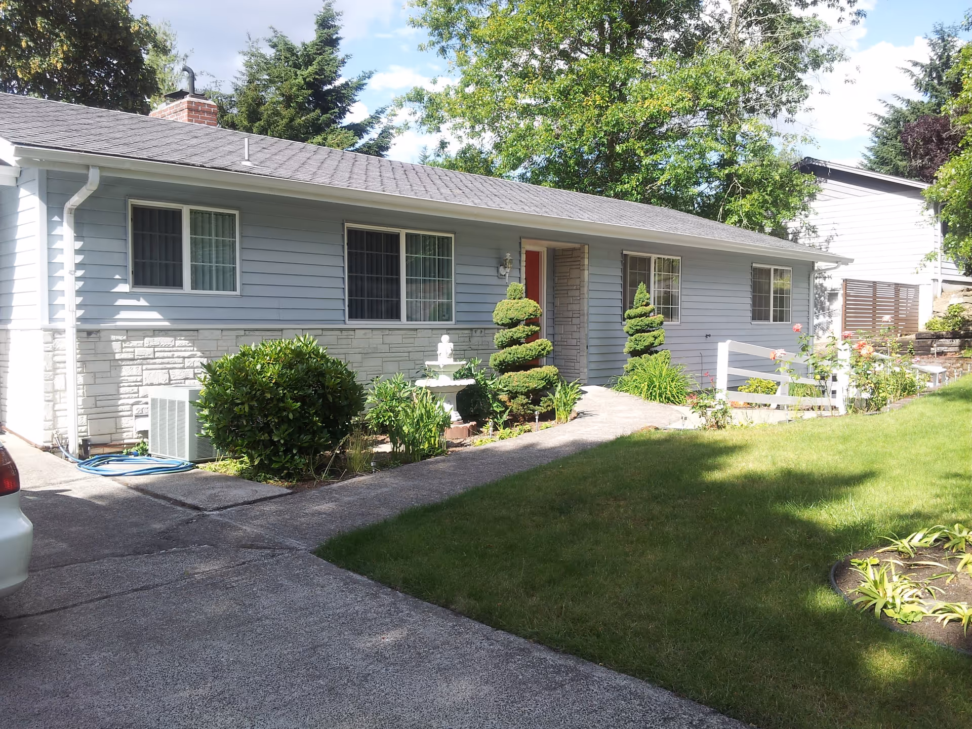 Single-story light-gray house front with a driveway, lawn, shrubs, and a walkway leading to the entrance.