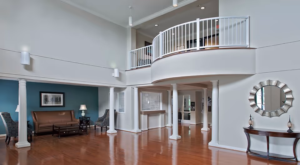 Spacious interior common area with polished wooden floors, white columns, and a curved balcony railing above. There is a seating area with a brown leather sofa, two patterned armchairs, a coffee table, and side tables with lamps against a teal accent wall. A decorative round mirror hangs above a wooden console table with two ornamental pieces on the right side of the image.